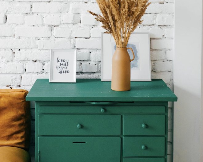 A green cabinet with a beige vase containing dried plants, placed next to two framed artworks against a white brick wall.