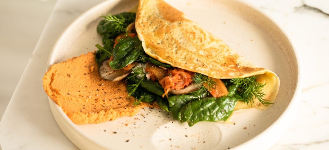 Golden-brown omelette filled with fresh spinach, herbs, and vegetables, served on a white plate with a side of sauce.